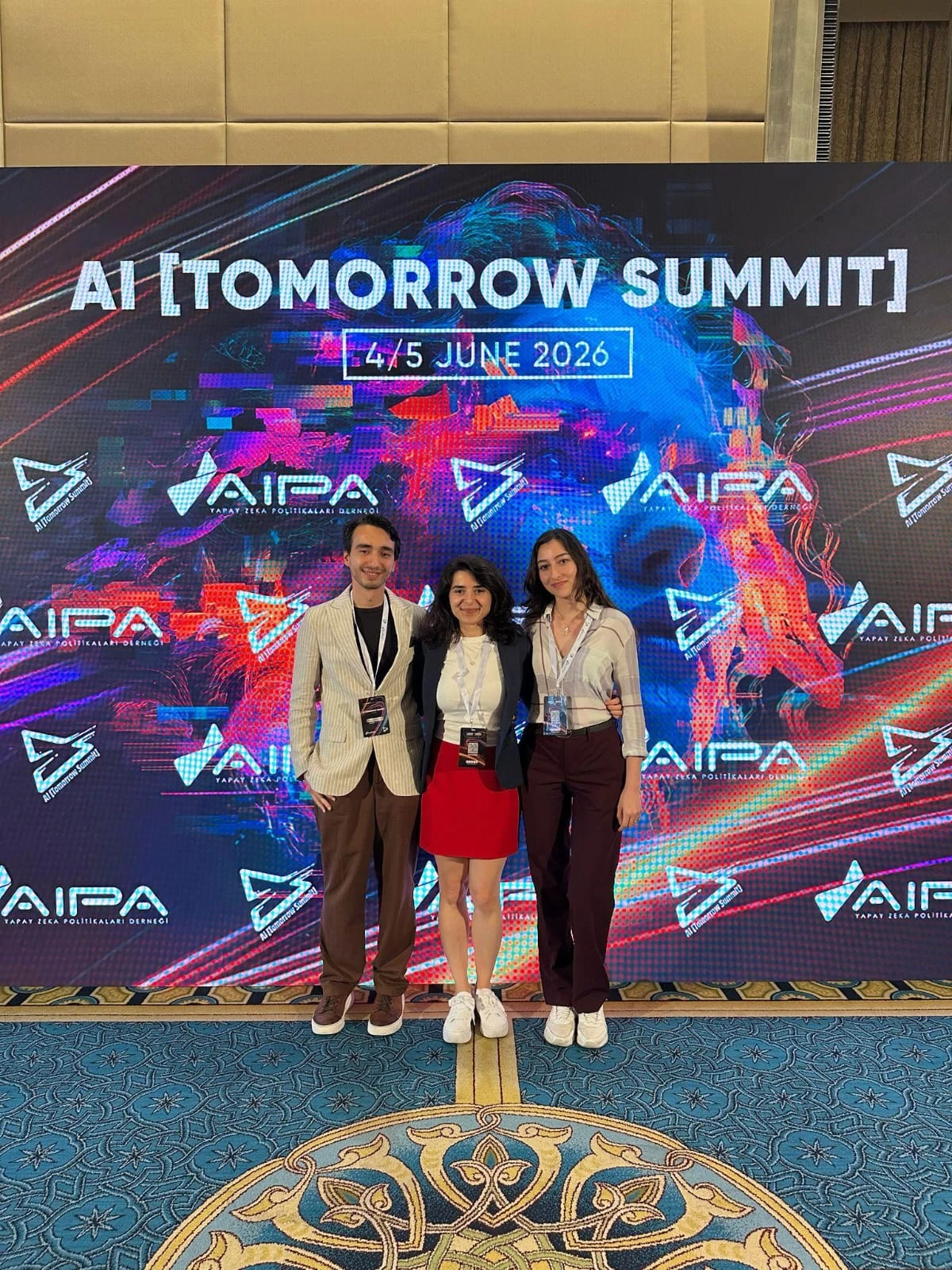 AI Tomorrow Summit ekranının önünde etkinlik kartlarıyla poz veren üç kişi